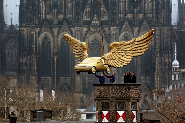 Keulen.- Workers install a golden winged car on the roof of the City Museum in Cologne, western Germany, on April 4, 2013. The car, a creation by German artist HA Schult, is brought back to the roof after its restauration at Ford. AFP PHOTO / OLIVER BERG.” (artdaily)